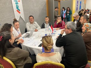 Yuvarlak bir masanın etrafında bir grup insan oturuyor. Masada çeşitli belgeler, bir dizüstü bilgisayar ve birkaç su şişesi bulunuyor. Masanın üzerinde Türk bayrağı ve "Yeşil Binalar" yazılı bir bayrak var. Katılımcılar arasında farklı yaş gruplarından insanlar var ve bazıları boyunlarında yaka kartları taşıyor. Arka planda, duvarda "Yeşil Binalar" yazılı bir afiş asılı. İnsanlar ciddi bir şekilde konuşuyor ve not alıyor gibi görünüyor. Arka planda başka insanlar da var, bazıları ayakta duruyor ve telefonla konuşuyor.