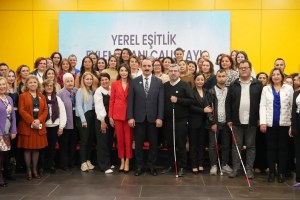 Kalabalık bir grup insan, bir etkinlikte bir araya gelmiş. Arka planda "Yerel Eşitlik Eylem Planı Çalıştayı" yazılı bir afiş var. Önde, takım elbise giymiş bir adam ve kırmızı takım elbise giymiş bir kadın dikkat çekiyor. Muhtemelen bu kişiler belediye başkanı veya belediye başkan yardımcıları olabilir. Çeşitli yaş ve cinsiyetten insanlar, gülümseyerek kameraya bakıyor. Bazı kişiler beyaz baston kullanıyor, bu da etkinliğin erişilebilirlik veya engellilikle ilgili bir temaya sahip olabileceğini düşündürüyor. Arka plan sarı renkte.