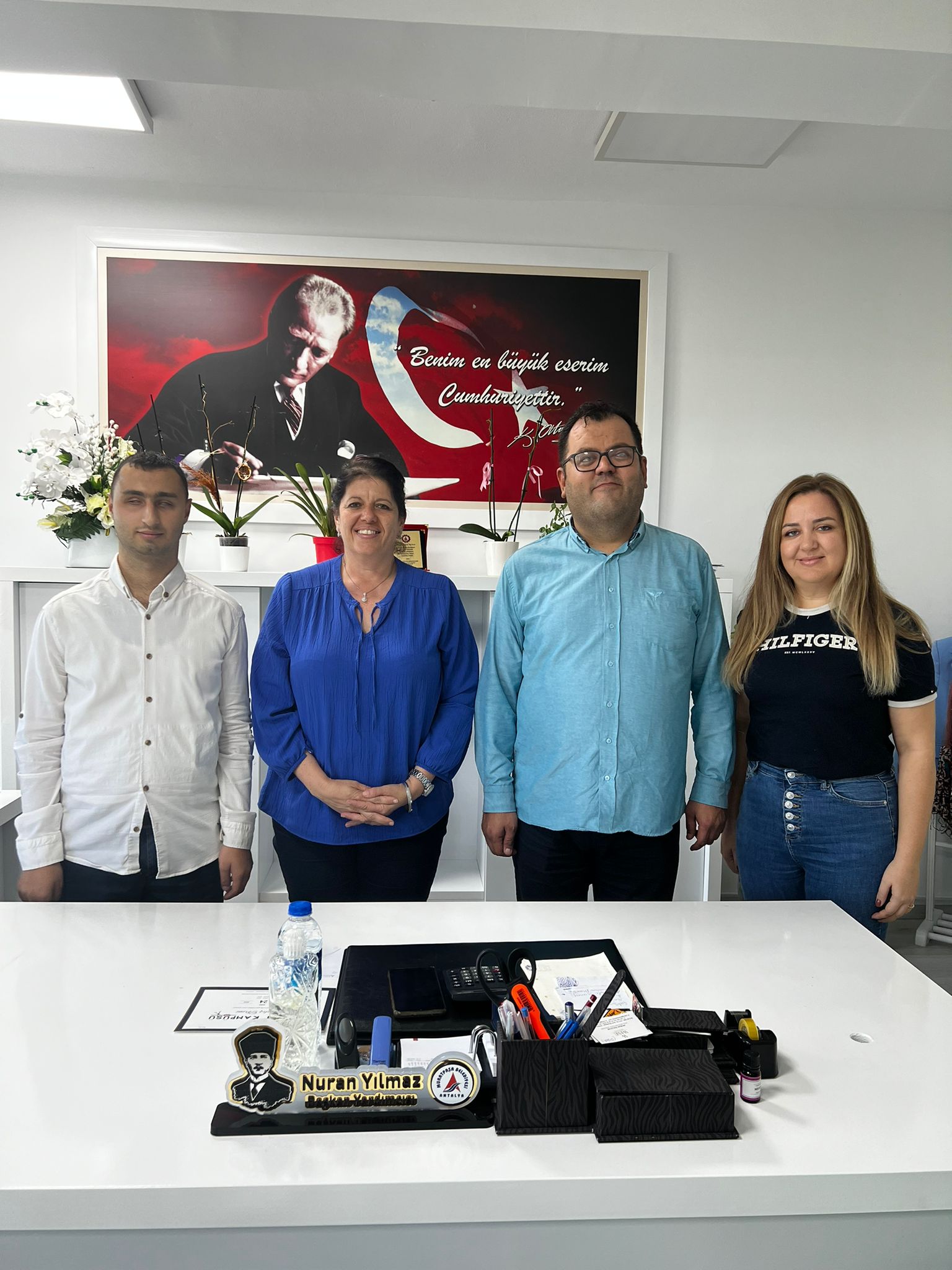 Fotoğrafta dört kişi bir masanın arkasında ayakta duruyor. Soldan sağa doğru, ilk kişi beyaz gömlek giymiş bir erkek. Yanında mavi bir bluz giymiş bir kadın var. Onun yanında açık mavi gömlek giymiş bir erkek duruyor. En sağda ise siyah tişört ve kot pantolon giymiş bir kadın var. Arkalarında, duvarda Atatürk'ün bir resmi ve